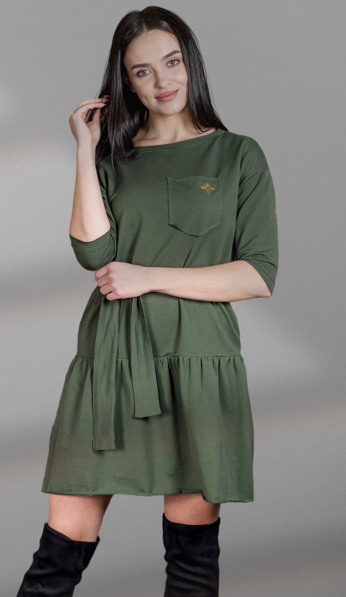 Modelka w oliwkowej sukience z kieszenią i haftem pszczoły, z rękawami 3/4 i paskiem, nosząca czarne kozaki za kolano, pozuje na szarym tle.
