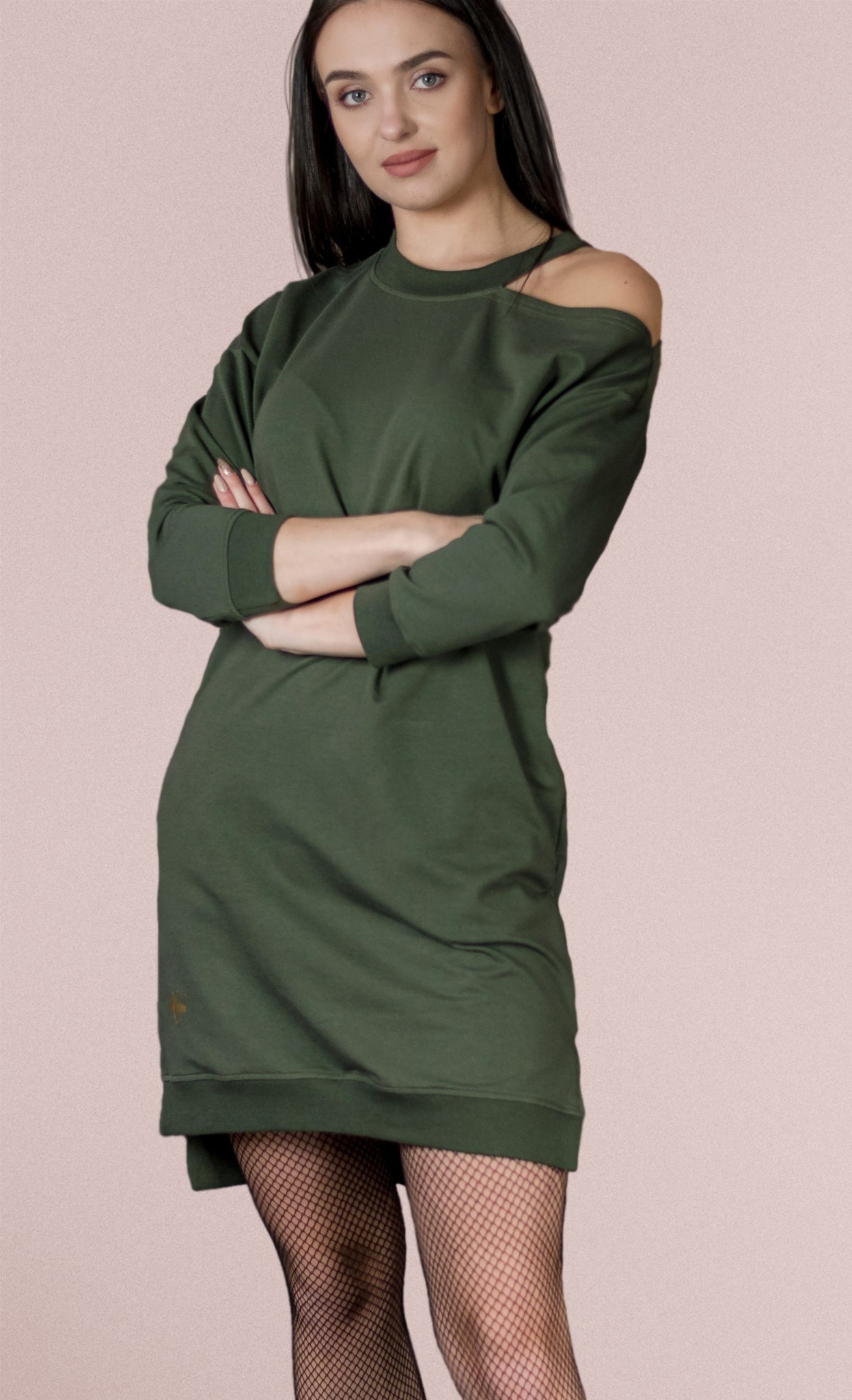Kobieta pozuje w oliwkowej sukience dresowej z odkrytym ramieniem i czarnych rajstopach kabaretkach na jednolitym tle.