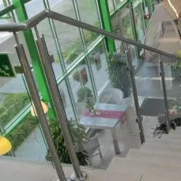Szklana balustrada z metalowymi poręczami na schodach w jasnym, nowoczesnym wnętrzu z dużą ilością zieleni i znakiem ewakuacyjnym.