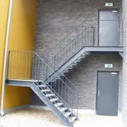 Zewnętrzne schody z metalu z szarymi poręczami, prowadzące do dwóch metalowych drzwi na różnych poziomach budynku o elewacji z szarej cegły.
