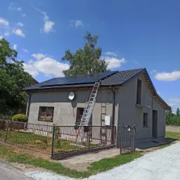 Dom jednorodzinny z zamontowanymi panelami słonecznymi na dachu, drabina oparta o ścianę, widok z zewnątrz w słoneczny dzień.