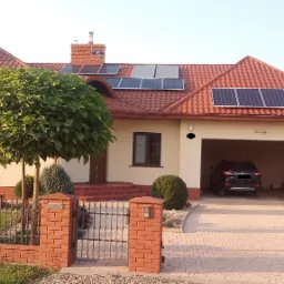 Dom jednorodzinny z czerwoną dachówką, na której zamontowane są panele słoneczne; garaż z samochodem w tle, ogrodzenie z cegły i kulisty krzew na pierwszym planie.