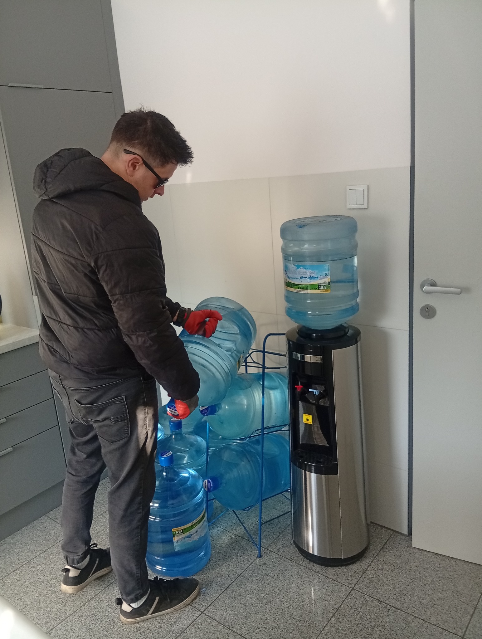Mężczyzna w rękawicach wymienia butlę z wodą w dystrybutorze. W tle zapas butli na metalowym stojaku. Widoczne elementy biura: szafki, drzwi.