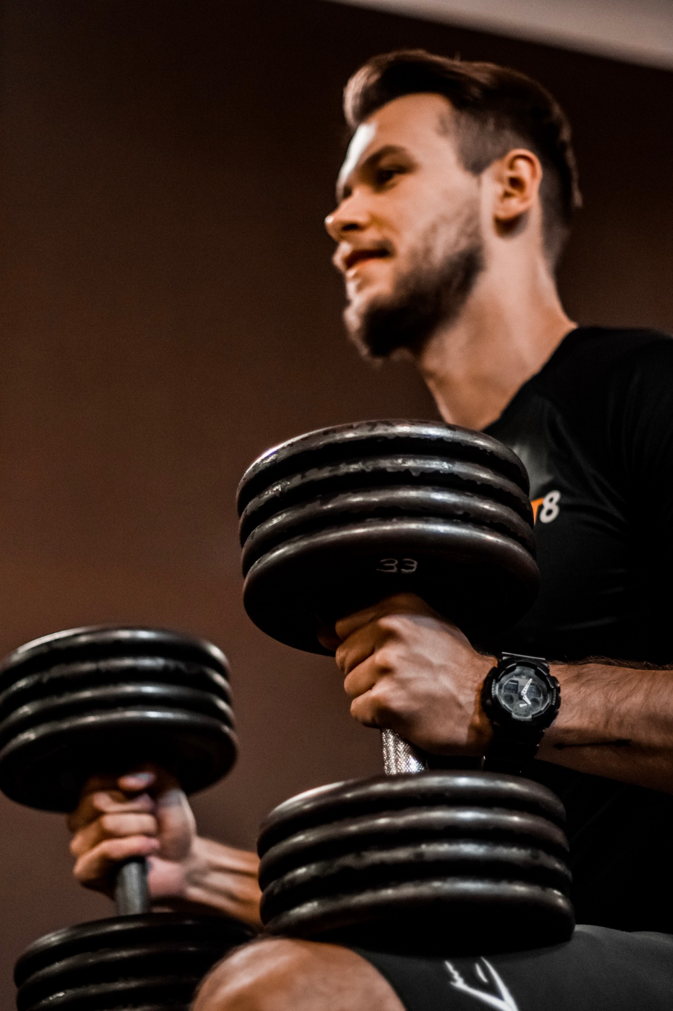 Mężczyzna w czarnej koszulce ćwiczy z dwoma ciężkimi hantlami, na nadgarstku widoczny czarny zegarek sportowy.