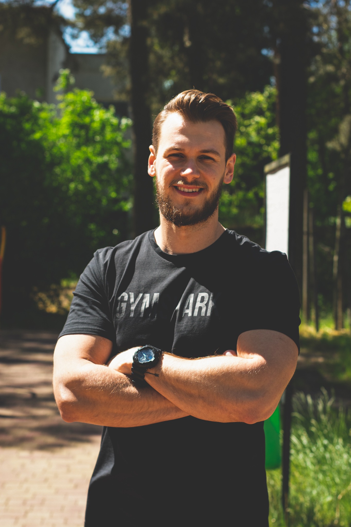 Uśmiechnięty mężczyzna z brodą, w czarnej koszulce z napisem GYM ARMY, stoi na zewnątrz z założonymi rękami, na nadgarstku widoczny sportowy zegarek.