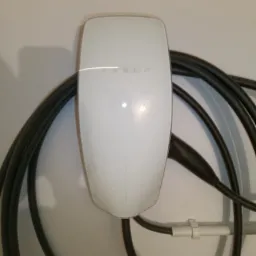 Biała stacja ładowania Tesla zamontowana na ścianie z widocznymi kablami, uchwycona z bliska.