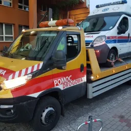 Żółto-czerwona laweta pomocy drogowej Iveco z włączonymi światłami przewozi białą karetkę pogotowia przed budynkiem szpitala.