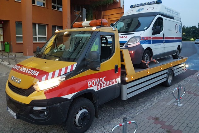 Żółto-czerwona laweta pomocy drogowej Iveco z włączonymi światłami przewozi białą karetkę pogotowia przed budynkiem szpitala.