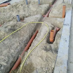 Wykop pod fundamenty z ułożonymi rurami kanalizacyjnymi i wentylacyjnymi, widoczne złącza i żółty przewód w piasku.
