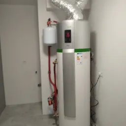 Biała pompa ciepła Haier z wyświetlaczem cyfrowym i rurami w pomieszczeniu z szarą podłogą z płytek.