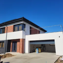 Nowoczesny dom z elementami cegły klinkierowej, białymi ścianami i otwartym garażem, widoczny teren wokół domu w trakcie prac wykończeniowych.