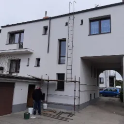 Malowanie elewacji domu dwupiętrowego na biało, widoczny pracownik z wiadrami farby, rusztowanie i drabina przymocowana do ściany, brama wjazdowa do garażu z boku.