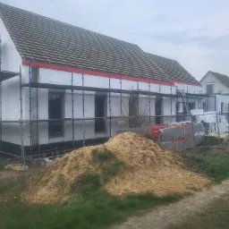 Budowa domu jednorodzinnego w trakcie ocieplania styropianem, widoczne rusztowania, przygotowane drewno na dach i sterta piasku.