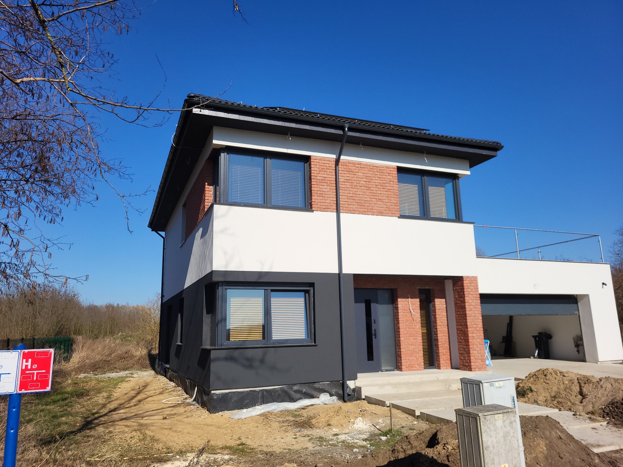 Nowoczesny, dwukondygnacyjny dom z elementami cegły klinkierowej na elewacji, pomalowany na biało i grafitowo, z widoczną izolacją przy gruncie i niezakończonym zagospodarowaniem terenu.