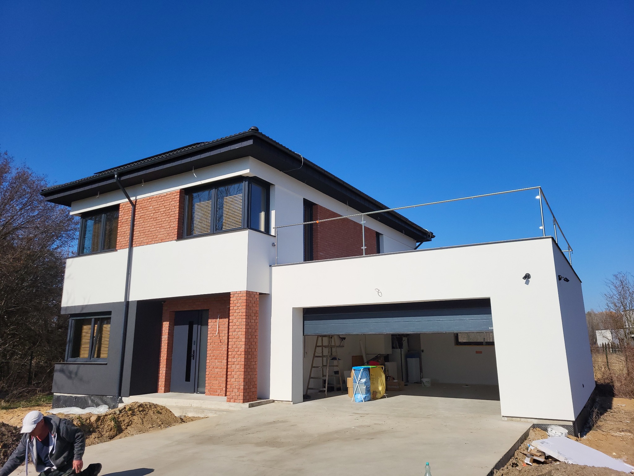Nowoczesny dom z elementami cegły klinkierowej, białymi ścianami i otwartym garażem, widoczny teren wokół domu w trakcie prac wykończeniowych.