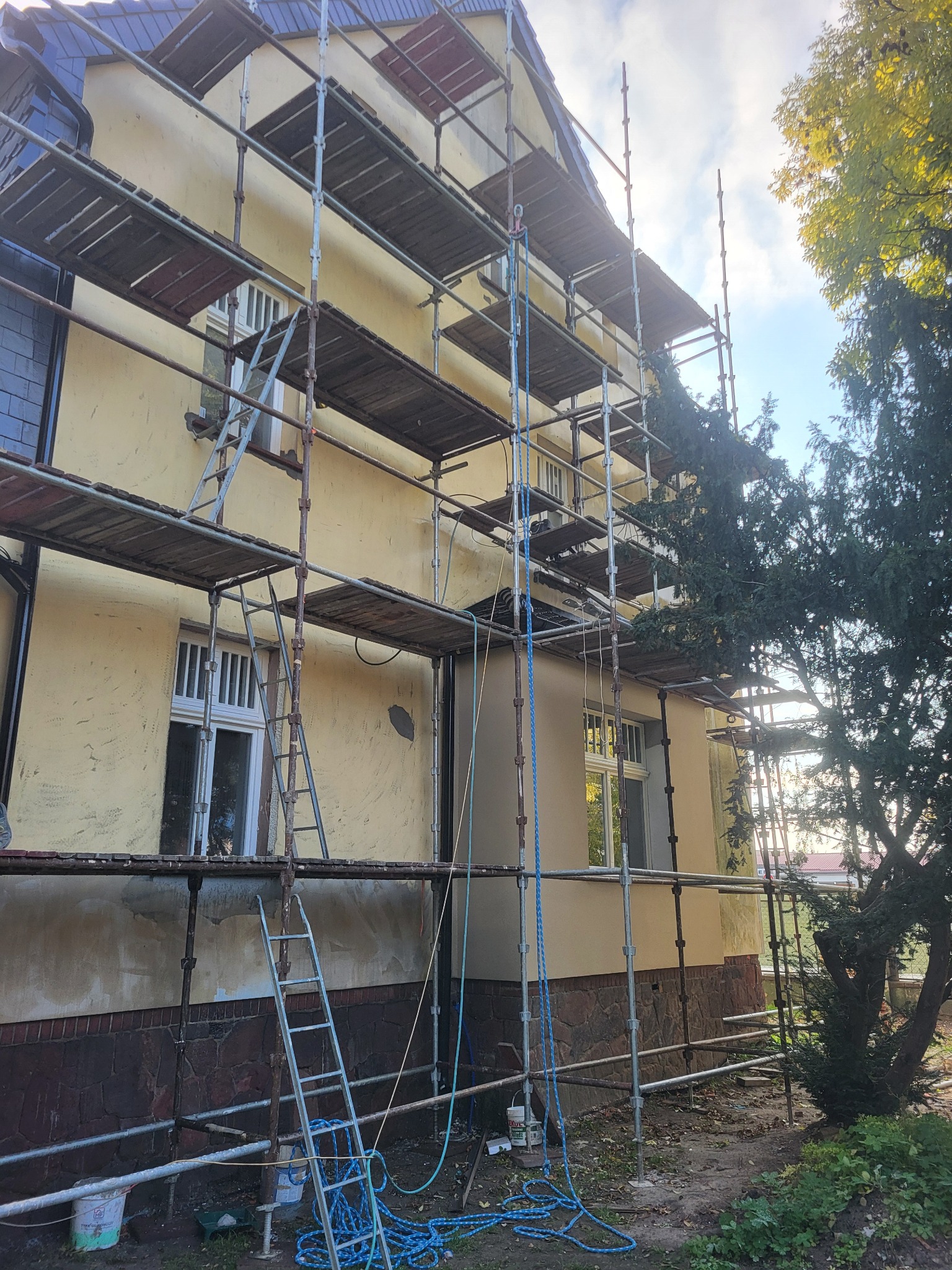 Dom w trakcie prac elewacyjnych, widoczne rusztowania, fragmenty starego i nowego tynku oraz wiadro z farbą u podstawy ściany.