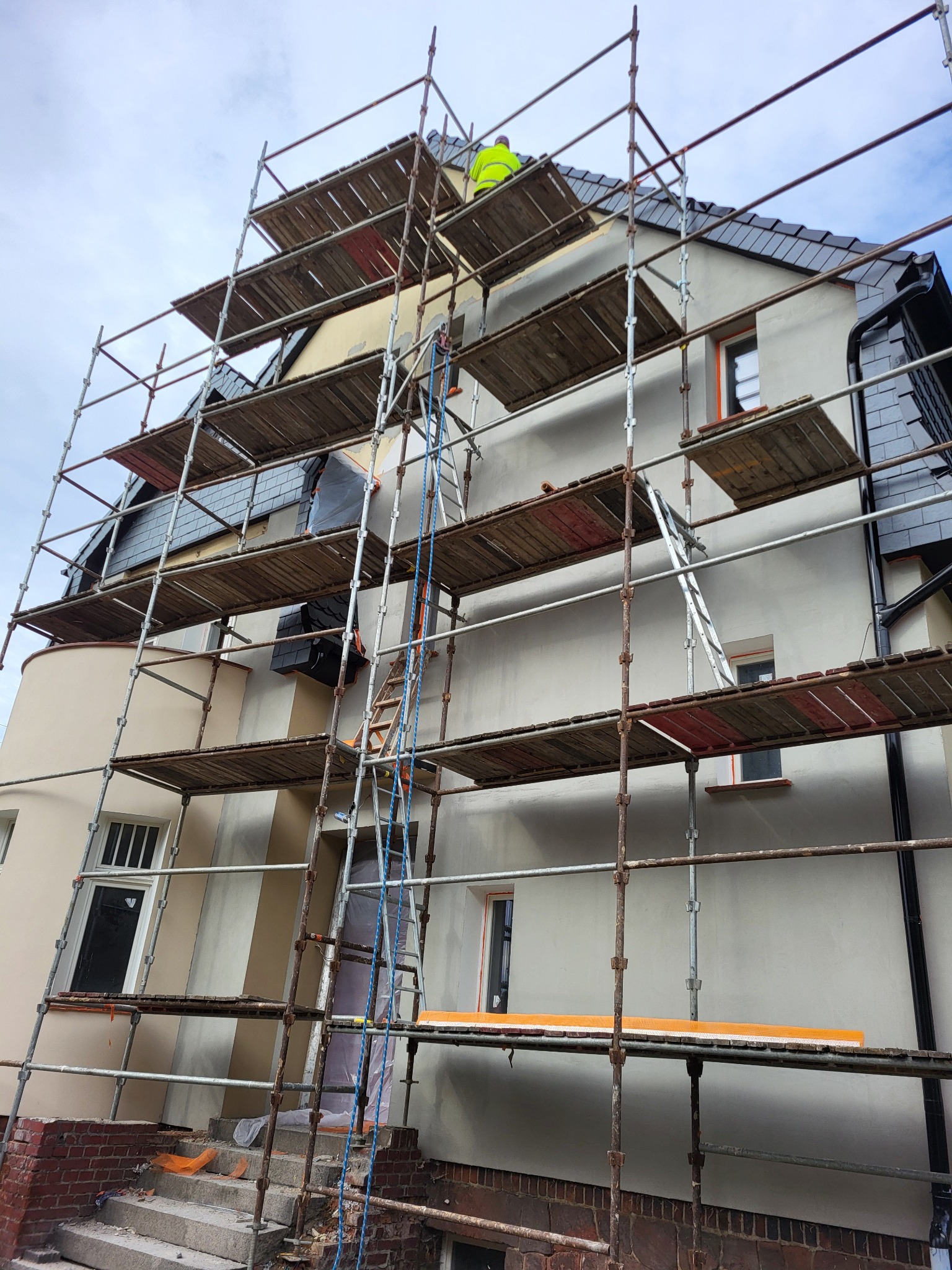 Dom w trakcie prac elewacyjnych, widoczne rusztowania, zabezpieczone wejście folią, robotnik w żółtej kamizelce na dachu, częściowo wykończona elewacja.