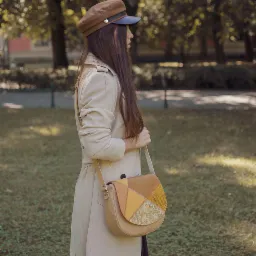 Kobieta w beżowym płaszczu i brązowej czapce prezentuje torebkę listonoszkę z patchworkowym wzorem na tle parku.