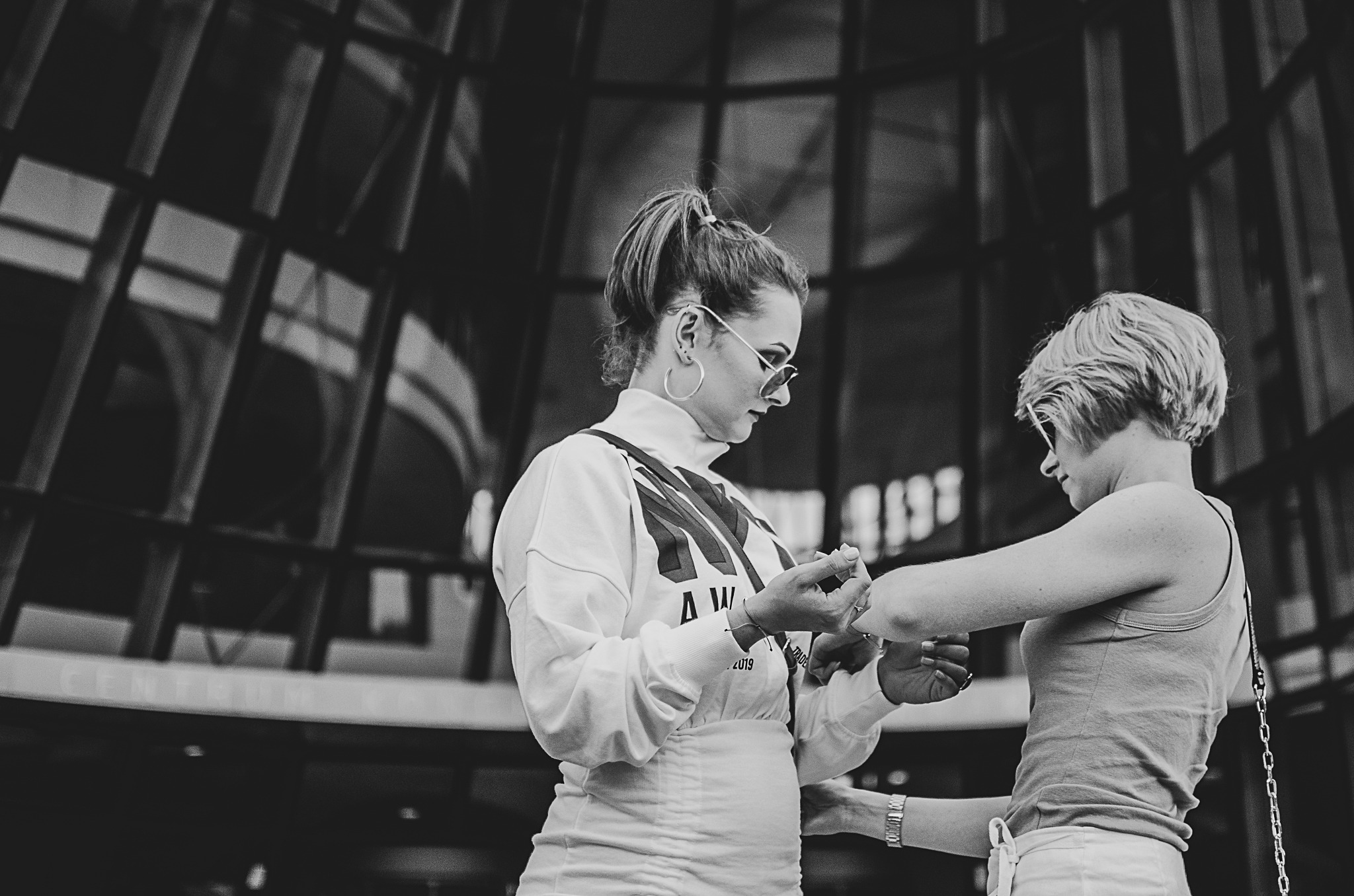 Czarno-białe zdjęcie dwóch kobiet w okularach, jedna z włosami upiętymi w kucyk, w trakcie stylizacji paznokci, na tle przeszklonej fasady budynku.