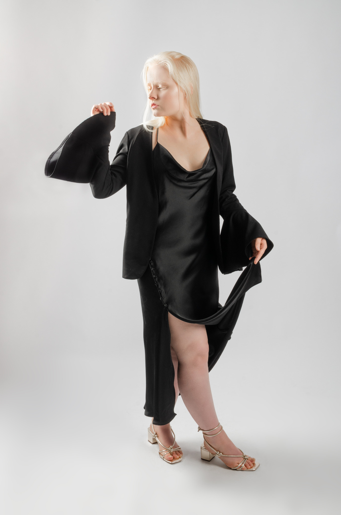 Modelka w czarnej satynowej sukience z dekoltem w kształcie wodospadu, czarnej marynarce z szerokimi rękawami i złotych sandałach na obcasie, pozująca na jasnym tle.