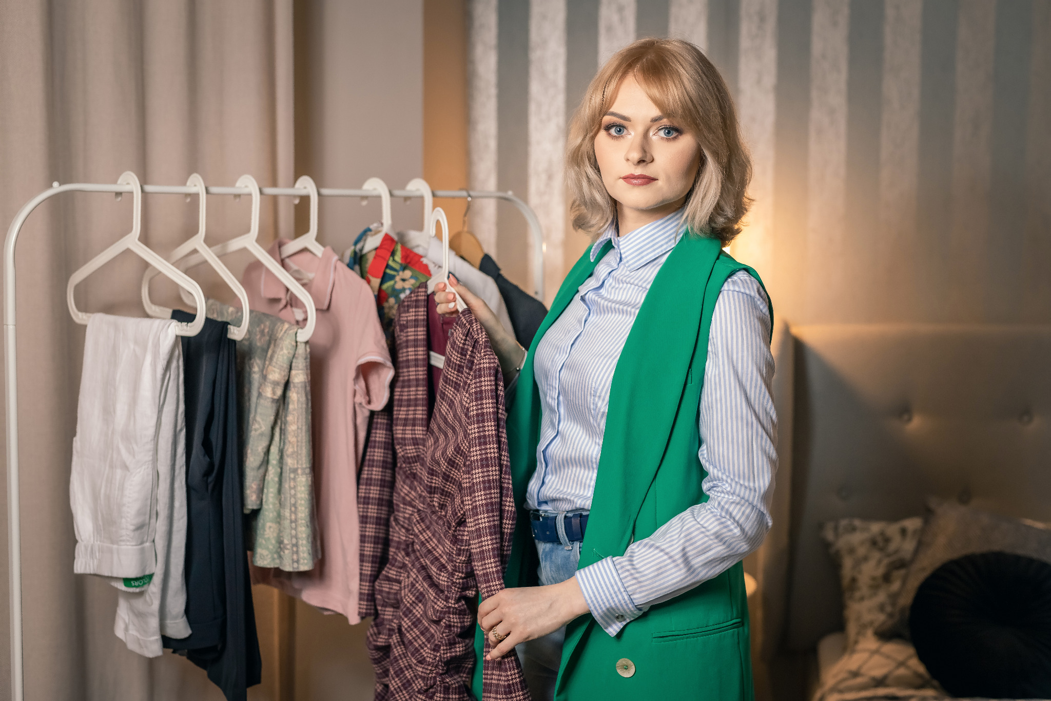 Kobieta w zielonej kamizelce i koszuli w paski, trzymająca w ręku kraciastą marynarkę na wieszaku, obok wieszaka z ubraniami w sypialni.