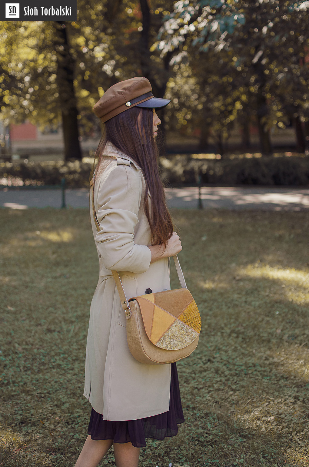 Kobieta w beżowym płaszczu i brązowej czapce prezentuje torebkę listonoszkę z patchworkowym wzorem na tle parku.