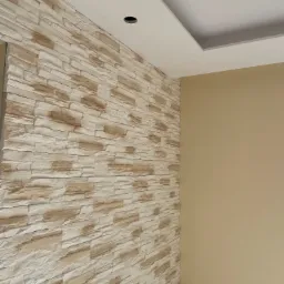 Ściana wykończona dekoracyjnym kamieniem w odcieniach beżu i bieli, sąsiadująca z gładką, pomalowaną ścianą w kolorze jasnego beżu. Sufit z podwieszanym elementem i otworem na oświetlenie.