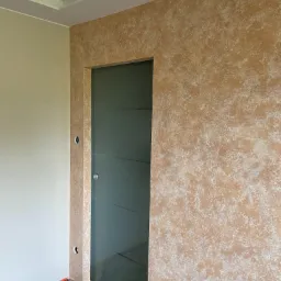 Ściana z efektem dekoracyjnym w odcieniach beżu i bieli, widoczna szklana ściana w ramie, podłoga zabezpieczona folią malarską, robocze obuwie przy ścianie.