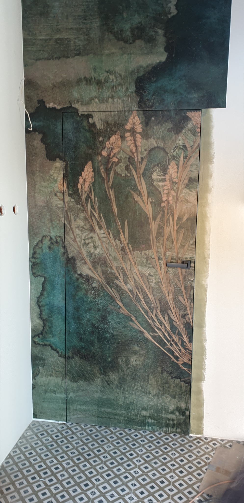 Ukryte drzwi w ścianie z motywem akwarelowych plam w odcieniach zieleni i brązu, ozdobione motywem roślinnym, widoczne wykończenie wokół ościeżnicy.