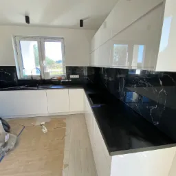 Nowoczesna kuchnia z białymi szafkami i czarnym blatem imitującym marmur, widoczne zabezpieczenia na podłodze podczas prac wykończeniowych.