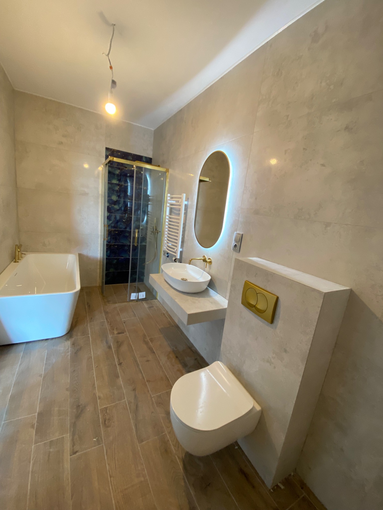 Nowoczesna łazienka z wanną, kabiną prysznicową z niebieskimi płytkami, podświetlanym lustrem i drewnopodobną podłogą, złote akcenty.