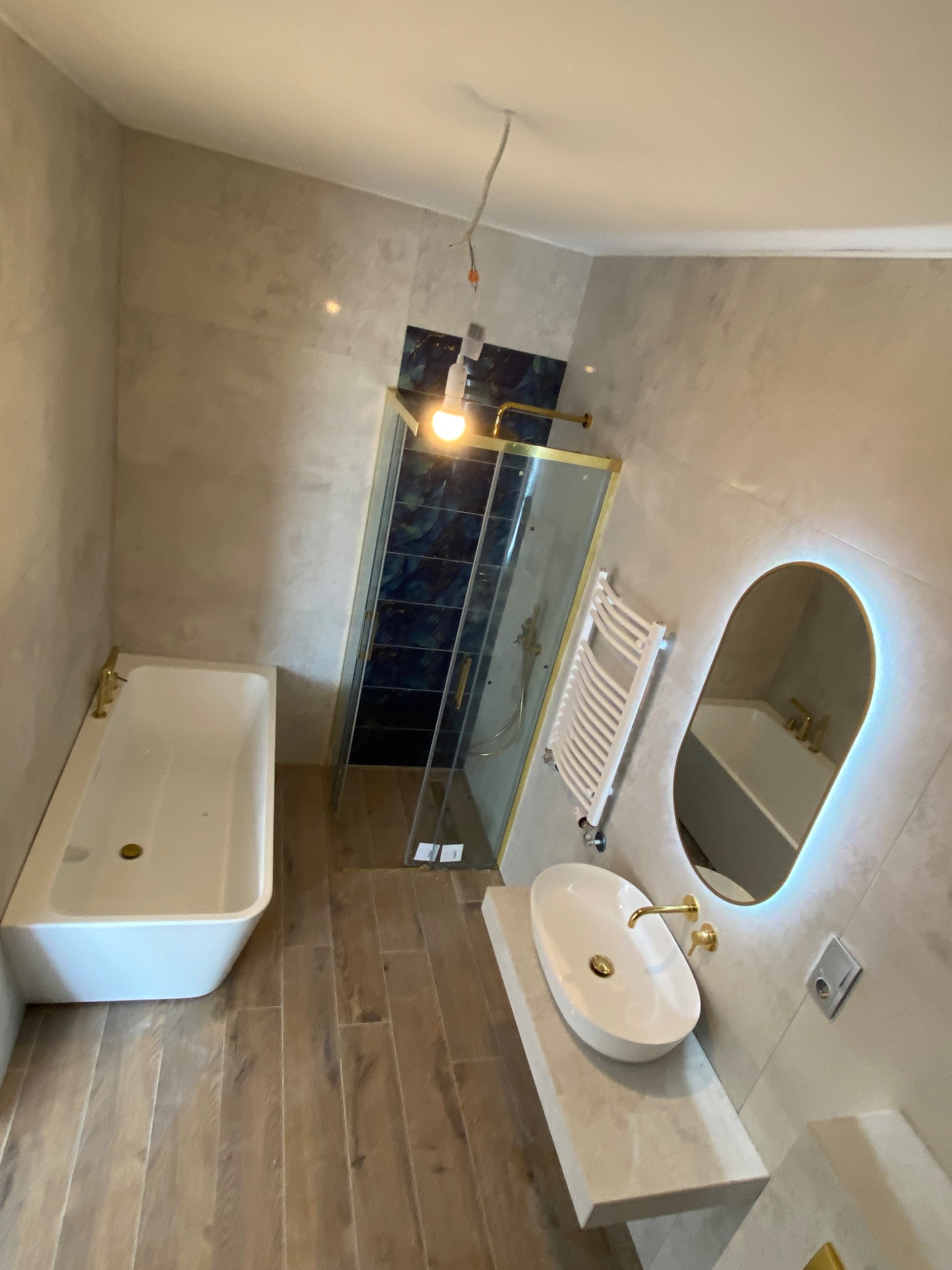 Nowoczesna łazienka w trakcie wykańczania, z wanną, kabiną prysznicową z ciemnymi płytkami, umywalką na blacie i podświetlanym lustrem owalnym, widoczne niedokończone podłączenia elektryczne.