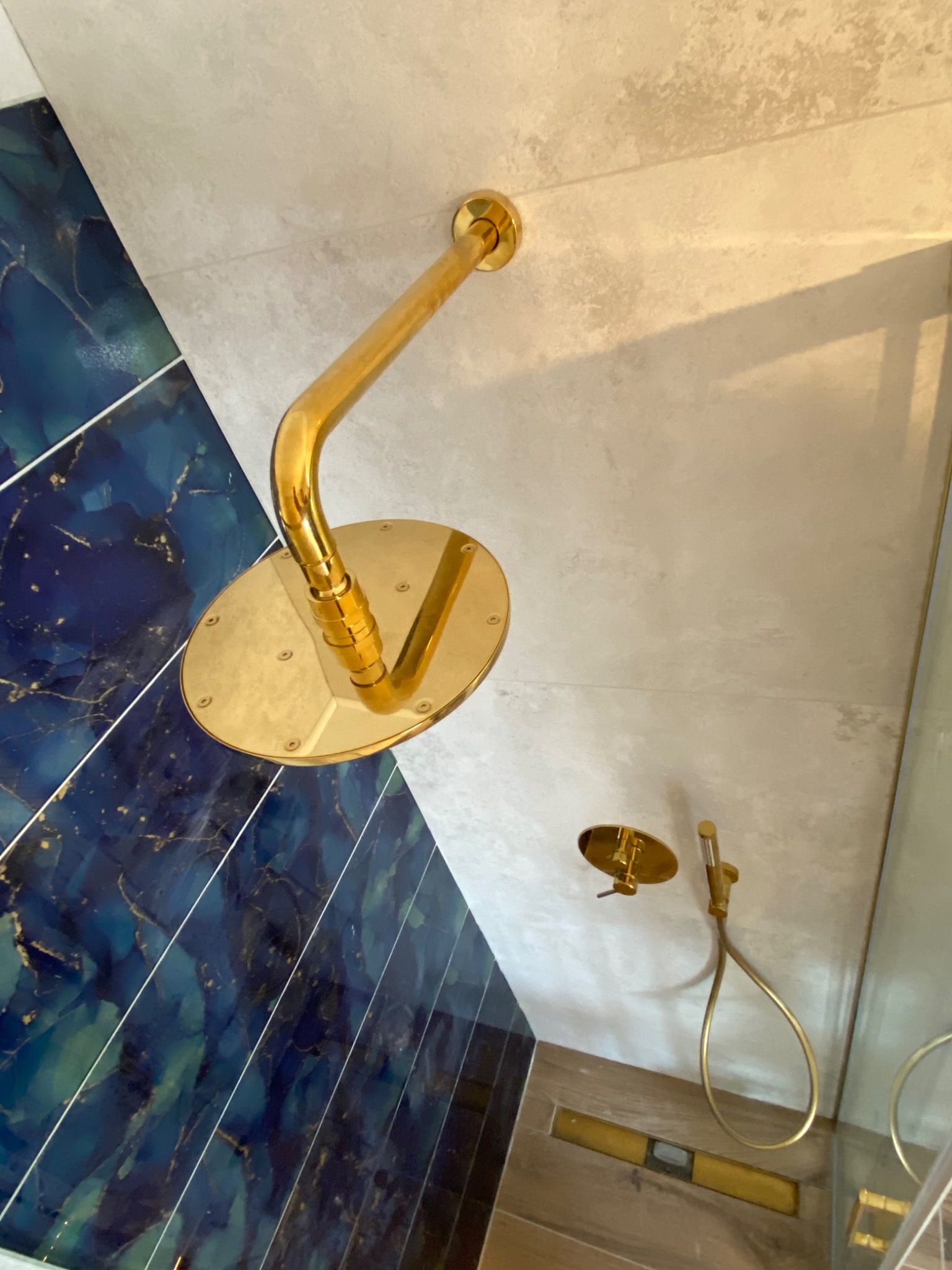 Złota deszczownica i bateria prysznicowa na tle płytek w odcieniach błękitu i szarości, widok z dołu.