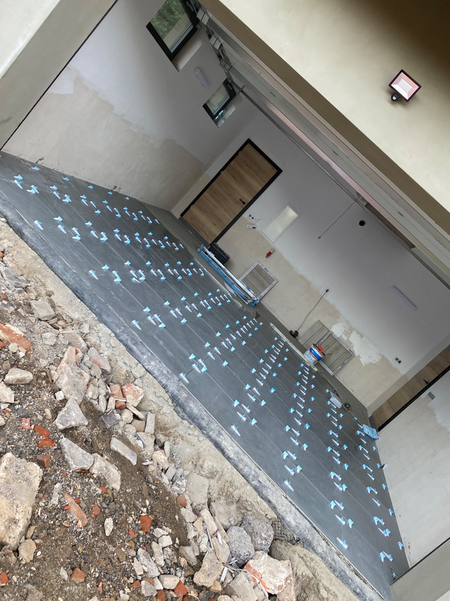 Układanie ciemnych paneli podłogowych z niebieskimi krzyżykami dystansowymi w trakcie remontu, widoczne gruz i niedokończone ściany.