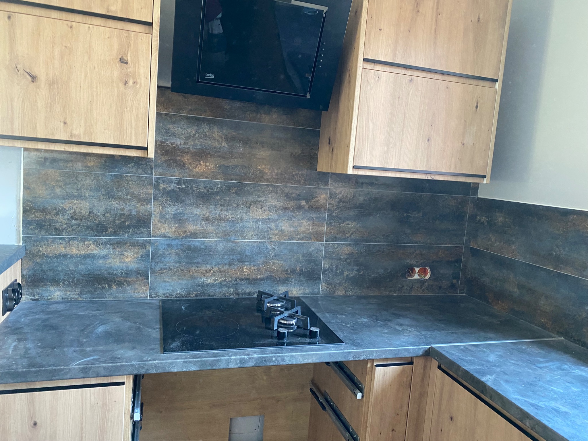 Nowoczesna kuchnia z drewnianymi szafkami o jasnym odcieniu, ciemnym blatem i płytkami imitującymi stal na ścianie, z czarnym okapem i płytą grzewczą z dwoma palnikami.