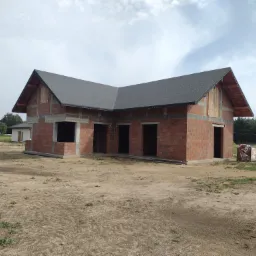 Surowy stan domu jednorodzinnego z cegły, z widocznym szarym dachem i otworami okiennymi, na piaszczystej działce w słoneczny dzień.