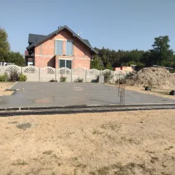 Wylany betonowy fundament z hydroizolacją i wystającymi prętami zbrojeniowymi, w tle budynek w trakcie budowy z czerwonej cegły i sterta ziemi.