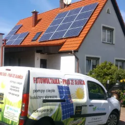 Dom jednorodzinny z panelami słonecznymi na pomarańczowym dachu, z przodu zaparkowany biały samochód dostawczy z reklamą firmy instalującej panele.