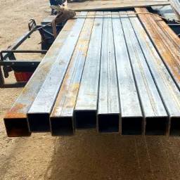 Kwadratowe profile stalowe, częściowo zardzewiałe, zabezpieczone pasami transportowymi na przyczepie samochodu dostawczego Renault Trafic.