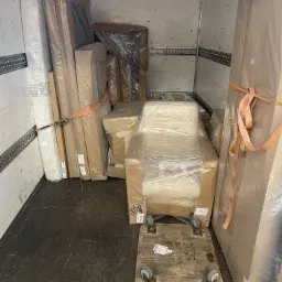 Wnętrze furgonetki wypełnione meblami zabezpieczonymi folią i kartonem, materac przywiązany pasem, krzesło na wózku transportowym.