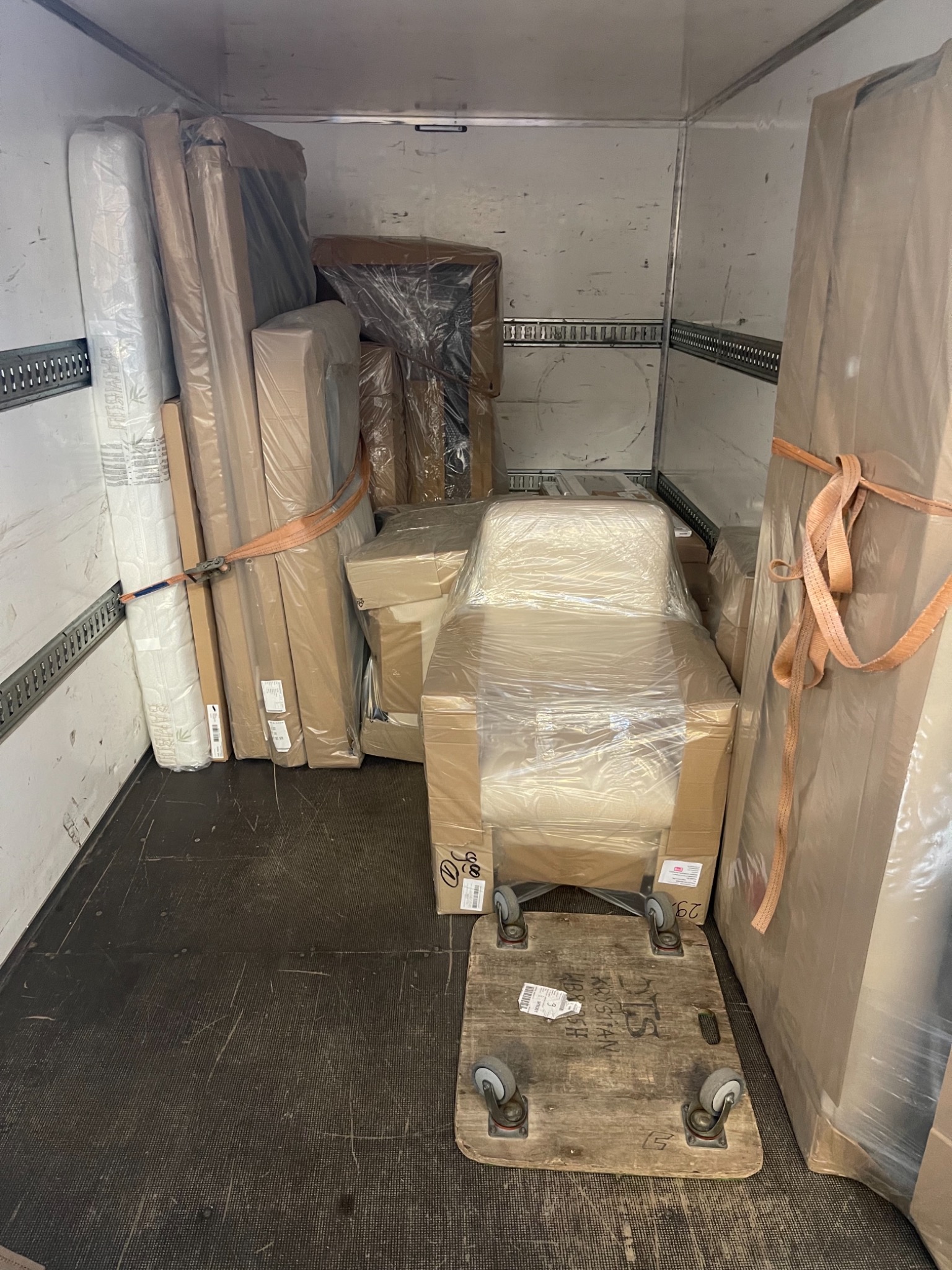 Wnętrze furgonetki wypełnione meblami zabezpieczonymi folią i kartonem, materac przywiązany pasem, krzesło na wózku transportowym.