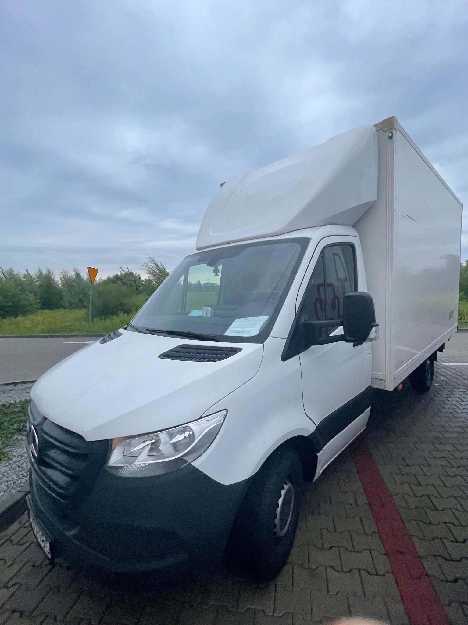 Biały samochód dostawczy typu kontener na parkingu z kostki brukowej, widoczny znak drogowy w tle, pochmurne niebo.