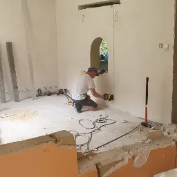 Pracownik w białej koszulce i niebieskiej czapce na kolanach wycina otwór w ścianie za pomocą piły elektrycznej. W tle widoczne resztki gruzu, narzędzia i betonowe elementy.