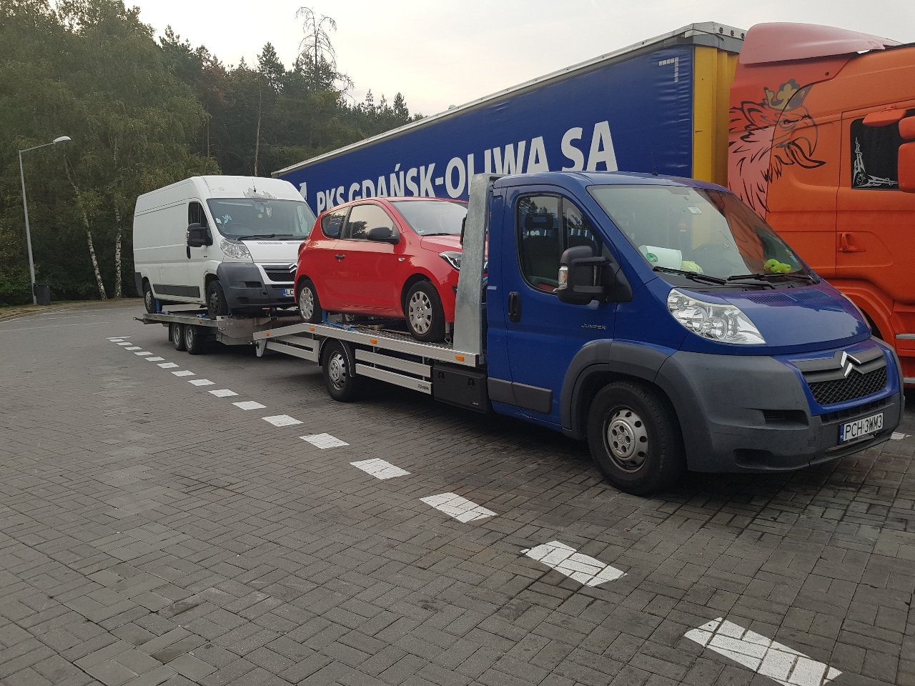 Niebieska laweta z czerwonym samochodem osobowym i białym busem na przyczepie, na parkingu z kostki brukowej, w tle ciężarówki i drzewa.