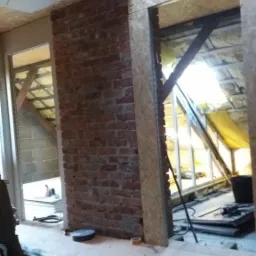 Surowa ściana z czerwonej cegły w trakcie budowy poddasza, widoczne drewniane elementy konstrukcyjne i izolacja, przewody elektryczne na podłodze.