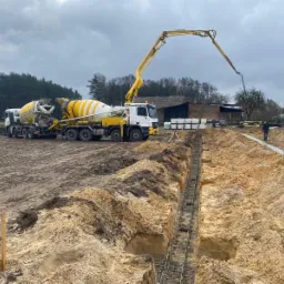 Żółto-biała betonomieszarka z wysięgnikiem wylewa beton do wykopu zbrojonego stalą pod przyszłe fundamenty domu jednorodzinnego na tle pochmurnego nieba i zabudowań gospodarczych.