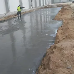 Wylewanie świeżej, gładkiej posadzki betonowej w hali przemysłowej, pracownik w kamizelce odblaskowej wygładza powierzchnię, widoczny fragment koparki w tle.