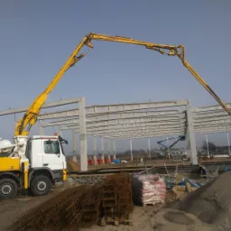 Żółto-biała betonomieszarka z wysięgnikiem pompuje mieszankę do szalunków pod konstrukcję hali, widoczne zbrojenia, worki z cementem, żuraw w tle.