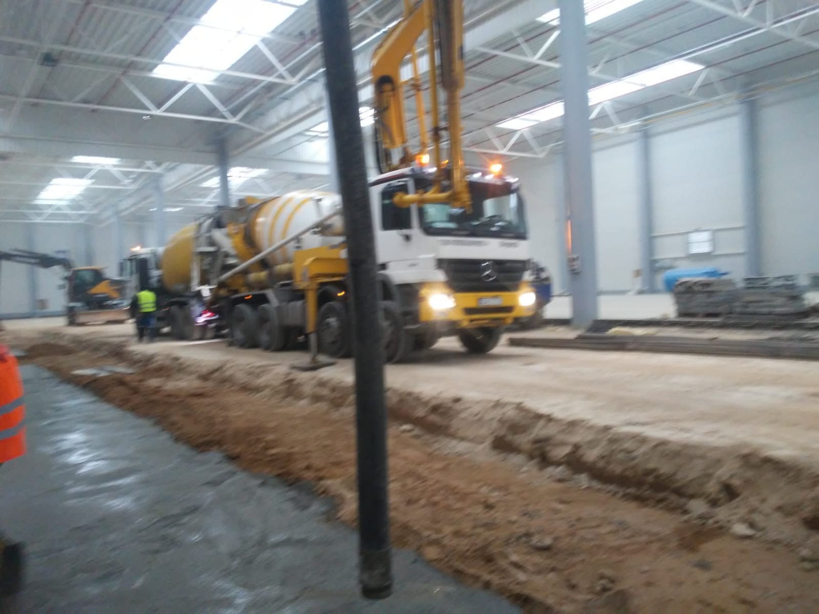 Wnętrze hali produkcyjnej z wylewką betonową w trakcie realizacji, widoczny żółto-biały betonomieszarka z wysięgnikiem oraz pracownicy w odblaskowych kamizelkach.