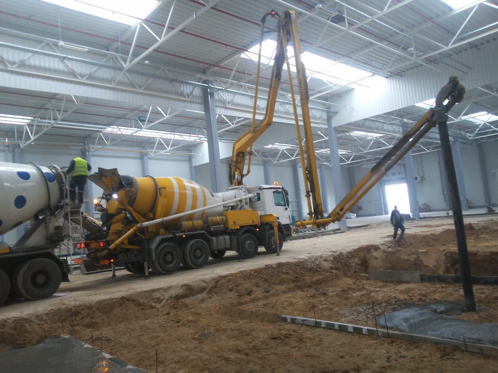 Wnętrze hali: żółto-biała betonomieszarka z pompą podaje beton do wykopu pod fundamenty, pracownik w żółtej kamizelce na drabinie, w tle widoczny inny pracownik.